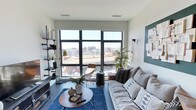 This single-floor space showcases a thoughtfully designed living area that blends modern aesthetics with comfortable, livable style. The room is anchored by a large, multi-pane floor-to-ceiling window that floods the interior with abundant natural light while offering expansive elevated views of the surrounding cityscape — a true standout feature of the space.

The living area is styled with a neutral palette of whites and grays, complemented by warm wood-toned accents. A plush, generously sized sofa adorned with patterned throw pillows faces the window, creating an inviting seating arrangement. A round wooden coffee table sits atop a deep blue area rug, adding a rich contrast to the light flooring.

Built-in open shelving along one wall provides functional display and storage space, while a teal-painted accent wall adds a bold, curated touch. A sleek pendant light and wall-mounted brass sconce contribute to the layered lighting design. A tall indoor plant introduces a natural element, softening the modern, urban atmosphere.

The overall ambiance is contemporary and airy, with clean lines and carefully selected furnishings that make the most of the elevated city setting.