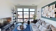 This single-floor space showcases a thoughtfully designed living area that blends modern aesthetics with comfortable, livable style. The room is anchored by a large, multi-pane floor-to-ceiling window that floods the interior with abundant natural light while offering expansive elevated views of the surrounding cityscape — a true standout feature of the space.

The living area is styled with a neutral palette of whites and grays, complemented by warm wood-toned accents. A plush, generously sized sofa adorned with patterned throw pillows faces the window, creating an inviting seating arrangement. A round wooden coffee table sits atop a deep blue area rug, adding a rich contrast to the light flooring.

Built-in open shelving along one wall provides functional display and storage space, while a teal-painted accent wall adds a bold, curated touch. A sleek pendant light and wall-mounted brass sconce contribute to the layered lighting design. A tall indoor plant introduces a natural element, softening the modern, urban atmosphere.

The overall ambiance is contemporary and airy, with clean lines and carefully selected furnishings that make the most of the elevated city setting.