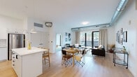 This thoughtfully designed single-floor residence showcases a contemporary aesthetic with clean lines and a bright, airy atmosphere. Exposed metallic ductwork runs throughout the space, lending an industrial character that contrasts attractively with the crisp white walls and light hardwood flooring found in the main living areas.

The open-concept layout seamlessly connects the kitchen, dining, and living areas. The kitchen is well-appointed with white cabinetry, subway tile backsplash, stainless steel appliances, a large island with seating, and pendant lighting overhead. The dining area comfortably accommodates a round table, while the living room benefits from generous natural light and direct access to a private outdoor balcony through floor-to-ceiling glass doors, offering views of the surrounding neighborhood and hillside scenery.

The bedrooms are carpeted for comfort, with one featuring its own balcony access and ample space. An additional room serves well as a home office or flexible space, complete with a built-in closet. Two full bathrooms feature white cabinetry, modern fixtures, and clean tile finishes—one with a tub-shower combo and one with a walk-in shower. The overall ambiance is modern, understated, and welcoming.