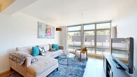 Located at 4040 26th Ave SW in Seattle's West Seattle neighborhood, this well-appointed one-bedroom, two-bathroom residence offers a thoughtfully designed single-level layout. The open-concept living and dining area is a standout feature, bathed in abundant natural light through expansive floor-to-ceiling windows that frame treetop and neighborhood views. Light hardwood-style flooring flows throughout the main living areas, adding warmth to the contemporary aesthetic.

The kitchen is outfitted with dark wood cabinetry, light countertops, and a full suite of stainless steel appliances including a refrigerator, range, microwave, and dishwasher. A kitchen island with bar seating provides additional prep space and a casual dining option.

The bedroom is a comfortable retreat featuring soft carpet, teal accent walls, a closet, and a window with an outdoor view. It connects conveniently to one of the two bathrooms, which includes a vanity with wood-toned cabinetry. The second bathroom features a full soaking tub and shower combination, also finished with clean, modern fixtures and light wood-style flooring. Together, the two bathrooms offer excellent functionality for residents and guests alike.