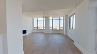 This single-floor unit is defined by its striking curved living area, where a continuous band of large floor-to-ceiling windows wraps along one entire wall, framing expansive panoramic city views from a notable elevation. The open layout is finished with light-toned hardwood flooring throughout, crisp white walls, and a ceiling fan overhead. A sleek fireplace anchors one wall of the living space, adding a focal point to the otherwise airy, minimalist design.

The kitchen features white flat-front cabinetry, open shelving, and white quartz countertops. Stainless steel and black appliances — including a range, microwave, and dishwasher — are complemented by a matte black undermount sink and matching hardware, creating a cohesive, contemporary aesthetic. Natural light enters through a window above the sink area.

The bathroom is finished in clean white subway tile and includes a soaking tub with matte black fixtures and a frosted window for natural light. The vanity area features a round mirror, white cabinetry with black hardware, and a white quartz countertop. Adjacent to the vanity, a walk-in closet provides generous storage with built-in shelving and hanging rods.