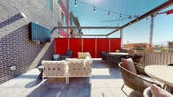 This rooftop terrace is a well-appointed outdoor amenity space designed for both relaxation and social gatherings. The terrace is finished with large-format porcelain tiles underfoot and is defined by striking bold red privacy panels that lend a modern, energetic character to the space while providing shelter from neighboring sightlines.

The terrace is organized into several distinct zones. A lounge area features generously cushioned sectional sofas and accent chairs arranged around a coffee table, ideal for casual conversation or unwinding. A separate dining zone accommodates a full-length wooden dining table with upholstered chairs, perfect for group meals. Additional intimate seating clusters with wicker and rope-woven chairs offer flexible arrangements for smaller gatherings.

A stainless steel outdoor grill station is positioned along the brick wall, providing a fully functional cooking setup to complement al fresco dining. An overhead pergola structure with retractable shade spans portions of the terrace, and strands of globe string lights are draped throughout, creating a warm, inviting ambiance into the evening hours.

Open sky views stretch across the surrounding urban landscape, with city rooftops and a construction crane visible in the distance, offering a dynamic backdrop. The combination of thoughtful furnishings, bold design accents, and practical amenities makes this rooftop terrace a versatile and welcoming outdoor destination.
