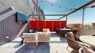 This rooftop terrace is a well-appointed outdoor amenity space designed for both relaxation and social gatherings. The terrace is finished with large-format porcelain tiles underfoot and is defined by striking bold red privacy panels that lend a modern, energetic character to the space while providing shelter from neighboring sightlines.

The terrace is organized into several distinct zones. A lounge area features generously cushioned sectional sofas and accent chairs arranged around a coffee table, ideal for casual conversation or unwinding. A separate dining zone accommodates a full-length wooden dining table with upholstered chairs, perfect for group meals. Additional intimate seating clusters with wicker and rope-woven chairs offer flexible arrangements for smaller gatherings.

A stainless steel outdoor grill station is positioned along the brick wall, providing a fully functional cooking setup to complement al fresco dining. An overhead pergola structure with retractable shade spans portions of the terrace, and strands of globe string lights are draped throughout, creating a warm, inviting ambiance into the evening hours.

Open sky views stretch across the surrounding urban landscape, with city rooftops and a construction crane visible in the distance, offering a dynamic backdrop. The combination of thoughtful furnishings, bold design accents, and practical amenities makes this rooftop terrace a versatile and welcoming outdoor destination.