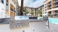 This outdoor amenity space is designed to offer a resort-style living experience within a multi-story residential community. At the heart of the area is a sparkling swimming pool, surrounded by lounge seating shaded by navy blue umbrellas, creating an inviting atmosphere for relaxation and recreation. Towering palm trees are thoughtfully integrated throughout the space, adding a tropical ambiance and natural shade.

The outdoor kitchen and grilling station is a standout feature, equipped with a stainless steel KitchenAid gas grill and elegant granite countertops set atop a stone-clad base, making it ideal for outdoor entertaining and communal gatherings. The structure is well-finished and built for durability in an open-air environment.

The surrounding multi-story building, with its warm golden and terracotta facade, red-tiled roofline, and numerous private balconies, frames the courtyard in a curved, embracing layout that enhances the sense of community. The architecture reflects a Mediterranean-inspired design aesthetic, complementing the lush landscaping below. Ample natural light fills the space, and the open-sky setting ensures a bright, airy feel throughout the day, making this amenity area a central and functional hub for outdoor living.