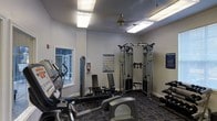 This fitness center in Denton, TX is a single-floor facility designed to support a well-rounded workout routine. The space is outfitted with a range of cardio machines, including treadmills and an elliptical, positioned to make efficient use of the floor area. A multi-station cable machine serves as the centerpiece for strength training, complemented by a weight bench and additional resistance equipment. The walls are painted in a clean, neutral gray, lending a bright and focused atmosphere to the room. Overhead fluorescent lighting ensures the space is well-illuminated throughout, while a ceiling fan provides added air circulation during workouts. A window along one wall allows for natural light to filter into the space. Instructional fitness posters are mounted on the walls, offering exercise guidance to users. The floor is covered in a durable, patterned carpet suited for a fitness environment. Informational signage is also posted, contributing to a structured and user-friendly setting. Overall, this fitness center provides a practical and functional environment with the essential equipment and amenities needed for a comprehensive workout experience.