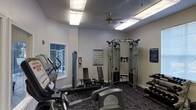 This fitness center in Denton, TX is a single-floor facility designed to support a well-rounded workout routine. The space is outfitted with a range of cardio machines, including treadmills and an elliptical, positioned to make efficient use of the floor area. A multi-station cable machine serves as the centerpiece for strength training, complemented by a weight bench and additional resistance equipment. The walls are painted in a clean, neutral gray, lending a bright and focused atmosphere to the room. Overhead fluorescent lighting ensures the space is well-illuminated throughout, while a ceiling fan provides added air circulation during workouts. A window along one wall allows for natural light to filter into the space. Instructional fitness posters are mounted on the walls, offering exercise guidance to users. The floor is covered in a durable, patterned carpet suited for a fitness environment. Informational signage is also posted, contributing to a structured and user-friendly setting. Overall, this fitness center provides a practical and functional environment with the essential equipment and amenities needed for a comprehensive workout experience.