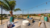 This expansive rooftop deck offers a compelling outdoor retreat elevated above the surrounding neighborhood, delivering sweeping panoramic views that stretch from the Pacific Ocean to the distant mountains and the palm-tree-lined cityscape below. The deck is finished with wide-plank composite wood flooring and is enclosed by a sleek cable-rail perimeter that preserves unobstructed sightlines from every angle.

The space is thoughtfully organized into multiple functional zones. A generously sized lounge area is anchored by a set of all-weather wicker sofas and armchairs in neutral tones, accented with blue and cream throw pillows, and centered around a round stone-finish coffee table. A separate dining area features a wooden table with bench seating, shaded by a vibrant chartreuse market umbrella. Chaise lounges provide a dedicated sunbathing area, ideal for relaxing under the California sun.

Tall, teal-glazed ceramic planters filled with bold flowering plants serve as striking decorative focal points throughout the deck, adding color and visual structure. A mature palm tree rises prominently from the space, providing natural shade and enhancing the tropical ambiance. Overhead, strands of Edison-style string lights crisscross the deck on black metal poles, creating a warm and inviting atmosphere for evening gatherings. Together, these elements make this rooftop deck an exceptional space for entertaining and relaxation.