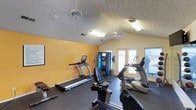 This single-floor fitness center offers a practical and functional space designed to support an active lifestyle. The room is finished with a textured ceiling and features vaulted rooflines that lend a sense of openness to the workout area. Warm yellow accent walls add an energizing atmosphere, complementing the dark rubber flooring that provides a safe and durable surface underfoot. The space is equipped with multiple cardio machines, including treadmills and a stationary recumbent bike, catering to a variety of fitness routines. A ceiling fan ensures comfortable air circulation during workouts, while fluorescent lighting provides consistent illumination throughout the room. A vending machine is conveniently positioned along the back wall, offering easy access to refreshments. A set of glass-panel French doors along one wall allows natural light to filter into the space and provides a direct connection to the outdoors. A wall-mounted display screen adds a modern touch and may serve as an entertainment or informational feature. The overall layout is open and uncluttered, maximizing usable floor space and allowing for easy movement between equipment. This fitness center is a straightforward, well-appointed facility suited for daily exercise routines.