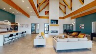 This single-story clubhouse is designed with a dramatic architectural presence, anchored by soaring vaulted ceilings adorned with striking exposed wood beam trusses. The open main gathering area centers around a floor-to-ceiling fireplace with a wall-mounted TV above, flanked by large black-framed glass doors that invite abundant natural light and provide direct views of the outdoor pool and patio. Comfortable neutral-toned seating arrangements create a welcoming lounge atmosphere throughout the space.

The fully equipped kitchen features rich wood cabinetry, a white tile backsplash, open floating shelves, and a bold teal accent wall that adds a vibrant contrast. Circular pendant lights hang above a large central island with bar seating, and stainless steel appliances, including a full-size refrigerator, complete the space. A dedicated game area includes a pool table, foosball table, and high-top tables positioned near large windows overlooking the pool courtyard. Additional seating alcoves and bar-height areas provide flexible gathering options. Stone-look tile flooring flows consistently throughout, unifying each zone of this well-appointed community clubhouse.