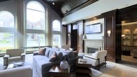 This stunning single-floor space makes an immediate impression with its dramatic vaulted ceiling, finished in dark wood paneling that creates a rich, layered contrast against the light-toned walls. The living area is anchored by a statement fireplace framed in classic stone and surrounded by floor-to-ceiling dark wood built-ins, complemented by wall sconces that add warm, ambient lighting. A wall-mounted flat-screen display is seamlessly integrated into the millwork above the fireplace, blending modern convenience with traditional craftsmanship. Towering arched windows flood the interior with abundant natural light, offering views of the surrounding neighborhood and lending an open, airy quality to the space despite its intimate layout. The seating arrangement — featuring a generous sectional sofa in cool grey tones accented with rust-colored throw pillows — pairs effortlessly with clean-lined white accent chairs, striking a balance between comfort and refined style. A dark wood coffee table with bold geometric detailing grounds the seating area over a patterned area rug. The overall ambiance is one of upscale elegance, where architectural detail, natural light, and carefully curated furnishings come together to create a welcoming yet sophisticated environment.