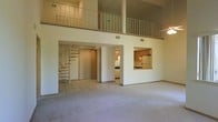 This two-story loft-style space offers an impressive sense of vertical volume, anchored by soaring double-height ceilings that draw the eye upward to an open upper-level loft with a metal railing balcony overlooking the main living area below. The expansive main floor is finished with plush carpet in the living area that transitions to tile flooring toward the back, creating a natural delineation between spaces. A striking spiral staircase with metal steps and a streamlined railing adds both architectural character and a space-efficient connection between floors. The kitchen is thoughtfully positioned with warm wood cabinetry and a pass-through window that opens directly into the living area, promoting an open and connected feel while maintaining a defined cooking zone. Standard appliances including a refrigerator and range are visible within the kitchen. A doorway adjacent to the kitchen leads to additional interior spaces. The upper level loft, accessible via the spiral staircase, provides a versatile secondary space ideal for a bedroom, office, or lounge area. A ceiling fan on the main floor adds a practical comfort element. The overall palette is neutral and bright, with white walls and light-toned finishes throughout that enhance the airy, open ambiance of this distinctive multi-level layout.
