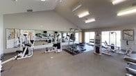 This fitness center is designed to support a comprehensive workout routine within a bright, open environment. The space features soaring vaulted ceilings that lend an airy, expansive feel throughout. A large wall-length mirror runs along one side of the main workout area, visually amplifying the space while allowing users to monitor their form during exercise.

The cardio section is outfitted with multiple treadmills and a stationary cycling bike, all positioned for easy use. Multiple flat-screen TVs are mounted on the walls, providing entertainment during workouts. The strength training area includes a well-rounded selection of cable machines, weight benches, free weights, and multi-functional strength stations, accommodating a wide range of exercises.

Flooring throughout the space transitions between carpet and rubberized mat sections, offering appropriate surfaces for both cardio and strength activities. Natural light filters in through large windows and a sliding glass door, contributing to the welcoming atmosphere. Additional amenities visible within the facility include a hand sanitizer station and an adjoining room accessible via a connecting doorway, suggesting supplementary facilities nearby. Overall, the fitness center is thoughtfully laid out to maximize usability and comfort for all levels of fitness enthusiasts.