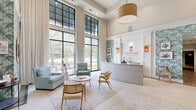 Located on Daniel Island in Charleston, South Carolina, this welcoming lobby space at Simmons Park serves as the leasing office and first impression for prospective residents. The single-floor layout is thoughtfully designed to balance function and style, featuring a dedicated reception desk prominently displaying the Simmons Park branding alongside warm pendant lantern lighting and curated botanical artwork.

The space is defined by its dramatic double-height ceilings and an expansive wall of floor-to-ceiling windows framed in dark green steel, allowing abundant natural light to flood the interior and offering unobstructed views of the lush outdoor greenery. Sheer ivory drapes soften the light while maintaining the airy, open atmosphere.

Tropical palm-print wallpaper in soft sage and teal tones lines the walls throughout, reinforcing a coastal Lowcountry aesthetic. The seating area is furnished with plush powder-blue armchairs, a round wood coffee table with hairpin legs, and a woven area rug, creating an inviting lounge atmosphere. Lush potted tropical plants add organic warmth throughout the space.

The entrance features bold dark green glass doors topped by transom windows, connecting visually to an exterior courtyard. A wall-mounted flat-screen display and additional seating nook complete the well-appointed interior, making this lobby both a functional and stylish gateway to the Simmons Park community.