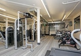 This modern fitness center is thoughtfully designed to support a full range of workout routines. The space is organized into distinct exercise zones, all unified by vibrant multi-colored striped carpeting that adds energy to the environment. Overhead, linear LED lighting runs throughout the ceiling, providing bright, even illumination across the entire floor.

The cardio zone lines one wall and features multiple treadmills, elliptical trainers, and a stationary spin bike, each equipped with individual digital displays. Wall-mounted flat-screen televisions are positioned above the cardio equipment, allowing users to stay entertained during their workouts. Rubber flooring mats are placed beneath the machines for added stability and floor protection.

The strength training area is anchored by a multi-station cable machine and includes a dedicated free weight section with a full rack of dumbbells, an adjustable weight bench, and additional resistance equipment. Structural columns define the space while large, motivational athletic photography decorates the walls, contributing to an energizing atmosphere.

Natural light enters through windows along the perimeter, and a set of large glass doors opens directly onto an outdoor patio furnished with seating, seamlessly connecting indoor and outdoor spaces. A water fountain is also conveniently located within the facility. The exposed ductwork overhead adds an industrial aesthetic that complements the clean, contemporary design of the space.