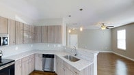 This well-appointed single-floor residence showcases a thoughtfully designed open-concept layout that seamlessly connects the kitchen and living areas. The kitchen is a standout feature, boasting light wood-toned cabinetry with sleek hardware, granite countertops with subtle speckled detailing, and a stainless steel undermount sink positioned at the peninsula. A stainless steel dishwasher adds a modern touch, while the peninsula itself creates a natural divide between the kitchen and the adjoining living space. Overhead, decorative pendant lights with a rustic mason jar aesthetic hang at varying heights, casting warm, inviting light over the workspace. The living area beyond benefits from a ceiling fan, ensuring year-round comfort. Neutral gray walls and light-toned flooring carry throughout, creating a cohesive and airy ambiance. The open layout allows for generous sightlines across the entire floor, enhancing the sense of space. A hallway visible in the background hints at additional rooms beyond, all tied together by the home's clean, contemporary aesthetic and light, neutral color palette throughout.