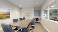 This professional conference room in Longview, Texas, is thoughtfully designed to support productive meetings and collaborative work. The room is anchored by a substantial rectangular conference table with a wood-tone finish and integrated cable management, surrounded by multiple high-back leather executive chairs that provide both comfort and a polished aesthetic.

The space features neutral-toned walls accented by carefully selected artwork, including a triptych of golden wheat field canvases and a large landscape print depicting a vibrant prairie sunset, adding warmth and character to the professional environment. Carpeted flooring in a subtle gray tone contributes to a quiet, focused atmosphere.

A dedicated workstation area along one wall includes blue-gray upper cabinetry and a built-in desk surface, offering additional functionality for administrative tasks or equipment placement. A pass-through window connects the conference room to an adjacent common area, enhancing connectivity within the broader office space.

Natural light enters through a window fitted with white horizontal blinds, allowing for adjustable illumination throughout the day. The drop ceiling incorporates recessed lighting and a standard tile grid, providing even and consistent overhead lighting. A private entry door with a glass panel further connects the room to the surrounding office environment, making this a well-equipped and versatile meeting space.
