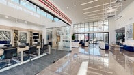 This single-floor amenity space is defined by its striking contemporary design and thoughtfully curated areas for work, wellness, and socializing. The expansive lobby welcomes visitors with polished marble-look tile flooring, soaring double-height ceilings, and floor-to-ceiling glass walls that flood the interior with natural light and offer views to the surrounding outdoor areas. A sleek glass-enclosed eLounge provides a dedicated workspace with workstations and seating, ideal for focused productivity. Adjacent to the lobby, an open lounge area features comfortable white and navy seating arrangements, large-format artwork, and oversized windows with black steel framing that reinforce the modern aesthetic. A full kitchenette serves the lounge with light wood cabinetry, white countertops, a stainless steel sink, beverage refrigerator, and snack storage. The fitness facilities are extensive, comprising a strength training room outfitted with a full cable machine system, free weights, benches, and floor-to-ceiling mirrors, as well as a cardio room with treadmills, ellipticals, and ceiling fans. A dedicated group fitness studio features vibrant striped wall graphics, wall-mounted TVs, TRX equipment, plyometric boxes, and rubber flooring, creating an energizing environment for group or individual training.