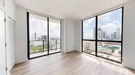 Located on the 19th floor at 1642 NE 1st Ave in Miami's vibrant Edgewater neighborhood, this one-bedroom, one-bathroom residence offers a modern, light-filled living experience with striking panoramic views of the Miami skyline and Biscayne Bay. The open-plan living area is defined by expansive floor-to-ceiling windows on two sides, flooding the space with natural light and framing breathtaking city and water vistas. Light-toned wood flooring runs throughout, complementing the clean white walls and creating a bright, contemporary aesthetic. A private balcony extends from the living area, offering unobstructed elevated views of downtown Miami's iconic skyline. The bedroom is a well-proportioned retreat featuring a full-height window with city views and a ceiling fan, while a louvered closet provides practical storage. The bathroom is finished in a clean, minimalist style with large-format neutral floor tiles, white subway tile in the walk-in shower, a floating vanity with a white countertop, and recessed lighting. A hallway connects the main living spaces efficiently. The unit also features a built-in kitchen area with appliances visible from the living space. The overall ambiance is sleek and modern, ideal for those seeking an urban Miami lifestyle with elevated views and refined finishes.