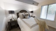 This single-floor residence offers a well-appointed interior with a refined aesthetic throughout. The bedroom serves as a standout feature of the space, showcasing thoughtful design and attention to detail. A distinctive angled soffit detail adds architectural character to the ceiling, creating visual depth and dimension in the room. Neutral tones in soft whites, warm grays, and champagne hues contribute to a calm and sophisticated ambiance. Natural light filters through a window fitted with horizontal blinds, brightening the space without overwhelming it. The room accommodates a generously sized bed with an elegant arched headboard in a rich dark finish, flanked by matching nightstands with dark-shaded table lamps that provide warm, ambient lighting. Plush carpeting underfoot adds a layer of comfort and warmth to the overall feel of the room. The carefully layered bedding, featuring textured pillows and a geometric-patterned throw, reflects a polished interior styling sensibility. The overall atmosphere is one of understated luxury — composed, inviting, and well-suited for rest and relaxation. The single-floor layout ensures ease of living, with the design elements working cohesively to create a harmonious and comfortable residential environment.