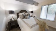 This single-floor residence offers a well-appointed interior with a refined aesthetic throughout. The bedroom serves as a standout feature of the space, showcasing thoughtful design and attention to detail. A distinctive angled soffit detail adds architectural character to the ceiling, creating visual depth and dimension in the room. Neutral tones in soft whites, warm grays, and champagne hues contribute to a calm and sophisticated ambiance. Natural light filters through a window fitted with horizontal blinds, brightening the space without overwhelming it. The room accommodates a generously sized bed with an elegant arched headboard in a rich dark finish, flanked by matching nightstands with dark-shaded table lamps that provide warm, ambient lighting. Plush carpeting underfoot adds a layer of comfort and warmth to the overall feel of the room. The carefully layered bedding, featuring textured pillows and a geometric-patterned throw, reflects a polished interior styling sensibility. The overall atmosphere is one of understated luxury — composed, inviting, and well-suited for rest and relaxation. The single-floor layout ensures ease of living, with the design elements working cohesively to create a harmonious and comfortable residential environment.