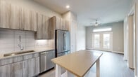Located at 2717 Minimax St in Houston's desirable 77008 zip code, this thoughtfully designed two-bedroom, one-bathroom unit offers a modern, open layout across a single floor. The kitchen is a standout feature, equipped with stainless steel appliances including a French door refrigerator, electric range, built-in microwave, and dishwasher. Driftwood-toned cabinetry extends to the ceiling, complemented by a white subway tile backsplash, light quartz countertops, and wood-look flooring that flows seamlessly throughout the main living areas. The living area is bright and airy, anchored by French doors topped with a transom window that open onto a private balcony, offering outdoor views and abundant natural light. A ceiling fan adds comfort to the space. Both bedrooms feature plush carpeting and closets with built-in shelving and hanging rods. The bathroom is finished with large-format gray tile surrounding a soaking tub and shower combination with chrome fixtures. Recessed lighting and a neutral color palette of warm whites and soft grays lend the entire unit a clean, contemporary ambiance well-suited for modern living.