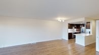 This two-storey space offers a bright, minimalist aesthetic defined by clean lines and a neutral palette throughout. The main floor showcases an open-concept layout where the living area flows seamlessly into the kitchen, creating a generous and versatile space ideal for both everyday living and entertaining. Light-toned hardwood-style flooring runs throughout, lending warmth and continuity to the interior.

The living area is expansive and unobstructed, with smooth white walls, white ceilings, and recessed floor vents that contribute to a tidy, streamlined appearance. A modern track light fixture provides focused overhead illumination, complementing the natural brightness of the space.

The kitchen is defined by rich, dark espresso cabinetry that creates a striking contrast against the lighter surroundings. It features stainless steel appliances, including a dishwasher, and a partial island or peninsula that subtly separates the kitchen from the living area while maintaining the open feel. Upper cabinets with under-cabinet lighting add both function and ambiance.

Spread across two floors, this space combines practical design with a contemporary finish, offering a comfortable and well-organized environment.
