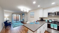 This single-floor unit is thoughtfully designed with a coastal aesthetic that carries throughout the space. The open-concept kitchen is a standout feature, outfitted with crisp white cabinetry, dark gray quartz countertops, and a full suite of stainless steel appliances, including a refrigerator, range, and over-the-range microwave. A peninsula island with bar-height seating provides a casual dining option and connects seamlessly to the living area, which is illuminated by a stylish multi-globe pendant light fixture. Warm hardwood-style flooring runs throughout the main living areas, complemented by a starfish-patterned teal area rug that reinforces the coastal theme. The living space opens directly to a private outdoor patio through a sliding glass door, extending the living area outdoors. A bedroom features plush carpeting, neutral walls, and a window with natural light. The bathroom offers a combined tub and shower, a vanity with a white countertop and gray cabinetry, and coordinating coastal-inspired décor. Throughout the unit, beach-themed wall art, soft teal and blue accents, and recessed lighting contribute to a bright, relaxed, and cohesive ambiance.