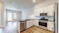 This well-designed one-bedroom, one-bathroom unit in Austin, TX offers a thoughtfully laid-out single-floor plan that maximizes every square foot. The open-concept kitchen and living area serves as the heart of the home, featuring crisp white shaker cabinetry, dark granite-style countertops, a subway tile backsplash, and a full suite of stainless steel appliances including a gas range, microwave, dishwasher, and refrigerator. Pendant lighting and recessed fixtures illuminate the space, while light wood-tone flooring flows throughout the main living areas, adding warmth and continuity. A ceiling fan overhead enhances comfort, and natural light pours in through blinds-equipped windows. The bedroom is a comfortable retreat finished with plush carpet flooring, a ceiling fan, and a window offering outdoor views. It is complemented by a dedicated walk-in closet with built-in shelving for ample storage. The bathroom features a soaking tub with a marble-style tiled surround, tile flooring, white cabinetry, and clean, neutral finishes throughout. The overall aesthetic is modern and cohesive, with a light, airy palette that creates an inviting and functional living environment.