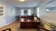 This single-floor office space serves as the leasing and administrative hub for Green Meadows Apartments. The layout is practical and professional, finished throughout with warm hardwood flooring and soft gray walls that create a calm, consistent aesthetic.

The entryway greets visitors with a classic six-panel white front door, a brass mail slot, and a branded welcome mat. A window fitted with horizontal blinds allows natural light to filter in, while a mounted fire extinguisher ensures safety compliance right at the entrance. A chair rail detail runs along the lower portion of the walls, adding a touch of architectural character.

The main office is generously proportioned, anchored by a large, polished wood executive desk with dual monitors and office equipment. Multiple windows on adjacent walls flood the room with natural light, and artwork adds a welcoming, professional touch. A secondary credenza and printer station complete the functional workspace.

Additional rooms branch off from the main office area, accessed through paneled white interior doors. One room features wall-mounted file organizers, offering organizational utility. Another smaller room is equipped with a window air conditioning unit flanked by potted palm plants, lending a refreshing, inviting feel.

Overall, the space is clean, functional, and thoughtfully arranged to support leasing operations in a comfortable, professional environment.