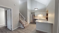 This two-story space offers a thoughtfully designed layout spread across two levels, characterized by a neutral color palette and wood-look flooring that flows throughout the main areas. The main level opens into a living area with soaring vaulted ceilings that lend an open, airy feel, complemented by a ceiling fan and a window fitted with blinds that allows natural light to filter in. A tiled fireplace with a white mantle serves as a prominent architectural feature in the living space.

The kitchen is well-appointed with white shaker-style cabinetry, light quartz countertops, a subway tile backsplash, and stainless steel appliances including a refrigerator and dishwasher. An extended breakfast bar provides additional counter space and casual seating. A pendant light fixture adds a finishing touch overhead. Carpeted stairs with white railings lead to the upper level, where additional living space features angled ceilings and windows. Two full bathrooms are present, each featuring white vanities with quartz countertops, tub-shower combinations, and wood-look tile flooring, maintaining the home's consistent, clean aesthetic throughout.