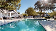 Located at 7816 Six Forks Rd in Raleigh, NC, this single-level outdoor amenity space is thoughtfully designed for relaxation and social gathering. At its center sits a expansive, sparkling swimming pool surrounded by a broad stone-paved deck lined with lounge chairs, offering ample space for sunbathing and leisure. A covered cabana anchors one end of the pool area, featuring elegant stone-clad columns, a beadboard ceiling, and a ceiling fan — providing a shaded retreat with direct views of the water. Adjacent to the pool, a white pergola structure offers a semi-private lounge area with built-in stone benches and comfortable seating, allowing filtered natural light to filter through its latticed panels. An elevated deck area provides elevated views of the surrounding tree-lined landscape, furnished with plush teal outdoor sofas ideal for casual conversation. A dedicated patio area houses an outdoor kitchen equipped with a built-in grill, complemented by a circular stone fire pit surrounded by Adirondack chairs — perfect for evening gatherings. Lush, mature trees frame the entire space, creating a serene, park-like atmosphere throughout.