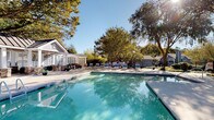 Located at 7816 Six Forks Rd in Raleigh, NC, this single-level outdoor amenity space is thoughtfully designed for relaxation and social gathering. At its center sits a expansive, sparkling swimming pool surrounded by a broad stone-paved deck lined with lounge chairs, offering ample space for sunbathing and leisure. A covered cabana anchors one end of the pool area, featuring elegant stone-clad columns, a beadboard ceiling, and a ceiling fan — providing a shaded retreat with direct views of the water. Adjacent to the pool, a white pergola structure offers a semi-private lounge area with built-in stone benches and comfortable seating, allowing filtered natural light to filter through its latticed panels. An elevated deck area provides elevated views of the surrounding tree-lined landscape, furnished with plush teal outdoor sofas ideal for casual conversation. A dedicated patio area houses an outdoor kitchen equipped with a built-in grill, complemented by a circular stone fire pit surrounded by Adirondack chairs — perfect for evening gatherings. Lush, mature trees frame the entire space, creating a serene, park-like atmosphere throughout.