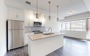 This one-bedroom, one-bathroom unit at 10043 108 Street NW presents a thoughtfully designed layout that maximizes its compact footprint. The open-concept kitchen and living area serves as the heart of the unit, featuring dark hardwood-style flooring that contrasts sharply with the crisp white walls and cabinetry. The kitchen is well-appointed with stainless steel appliances, including a full-size refrigerator, electric range, and built-in microwave, all complemented by a large central island with an undermount sink and quartz countertop. Stylish gold-toned pendant lights hang above the island, adding a contemporary accent. The living area connects seamlessly to expansive floor-to-ceiling windows and a sliding glass door, flooding the space with natural light and offering striking views of the downtown Edmonton skyline. The bedroom, accessed via a sliding door, is a cozy, well-defined space with hardwood-style flooring and direct access to a dedicated closet. The bathroom features light tile flooring, a full soaker tub with an overhead shower, a modern vanity with under-cabinet storage, and a wall-mounted mirror. A clean hallway with overhead ventilation connects all areas efficiently, reinforcing the unit's modern, functional design.