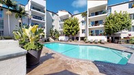 This outdoor amenity area is designed to deliver a resort-like experience, centered around a generously sized swimming pool with clear blue water. The pool is framed by a broad stamped-concrete sundeck finished in warm earth tones, providing ample space for lounging with modern reclining chairs positioned throughout. The surrounding multi-story building displays a Spanish-influenced architectural style, characterized by white stucco exteriors, red tile roof accents, and private balconies that overlook the courtyard below.

Lush tropical landscaping enhances the atmosphere, with mature palm trees, banana plants, flowering bromeliads, and an assortment of tropical shrubs arranged in decorative planters and raised garden beds. The greenery creates a sense of privacy and seclusion while adding vibrant color to the space.

A dedicated outdoor lounge and dining area is positioned poolside, furnished with modern patio seating and shaded by large market umbrellas. An outdoor kitchen station, equipped with a built-in stainless steel gas grill and stone-finished counter, provides a convenient space for al fresco cooking and entertaining. The overall ambiance is bright, open, and sun-drenched, consistent with a Southern California climate.