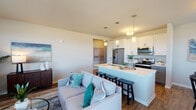This thoughtfully designed single-floor residence offers a seamless open-concept layout that connects the living, dining, and kitchen areas into one cohesive and functional living space. The kitchen is a standout feature, outfitted with crisp white upper cabinetry, gray lower cabinetry, a suite of stainless steel appliances including a French door refrigerator, electric range, microwave, and dishwasher, and a light-toned countertop island with bar seating. Pendant lighting above the island adds a refined touch to the space. The living area is bright and welcoming, anchored by large windows that invite generous natural light and offer views of the surrounding landscape. A dedicated dining area also benefits from oversized windows framing outdoor views, creating an airy and pleasant atmosphere for everyday meals. The primary bedroom is a comfortable retreat, complete with a walk-in closet and direct access to a private en-suite bathroom featuring gray cabinetry, a white countertop vanity, and wood-look flooring. A second bedroom provides additional flexibility and also includes a walk-in closet and natural light from a large window. The second full bathroom features a shower-tub combination and matching gray vanity. Warm wood-look flooring flows consistently throughout the main living areas, contributing to a cohesive and modern aesthetic throughout the home.