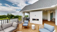 Located at 49 N Assembly Dr in Wilmington, NC, this elevated outdoor amenity space is thoughtfully designed for communal gathering and relaxation. The space is characterized by its warm herringbone-patterned brick flooring, crisp white shiplap and board-and-batten wall finishes, and a pitched gray shingle roof that ties seamlessly into the building's coastal architectural aesthetic.

The main covered lounge area serves as the heart of the space, anchored by a striking white tile fireplace with a mounted flat-screen television above, surrounded by comfortable seating arrangements that invite conversation and leisure. Ceiling fans provide airflow throughout the covered sections, enhancing year-round usability.

An outdoor kitchen is equipped with a full-size stainless steel grill and a stainless range hood, backed by a decorative tile surround — ideal for entertaining. Adjacent to the kitchen, a bar counter with high seating overlooks the outdoors through large navy-framed windows, blending indoor comfort with an open-air feel.

A dedicated covered dining area features a wood table and chairs alongside a wall-mounted television, perfect for al fresco meals. The space opens to expansive balcony areas with black metal railings that frame sweeping views of the surrounding greenery and community landscape.

A well-appointed bathroom with white subway tile walls, wood-look flooring, and accessible fixtures rounds out the amenities of this versatile outdoor retreat.
