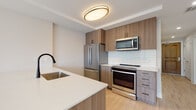 This thoughtfully designed single-floor unit combines modern aesthetics with functional living. The kitchen is a standout feature, showcasing warm wood-tone cabinetry that extends floor to ceiling, complemented by crisp white quartz countertops and a peninsula with an undermount sink fitted with a matte black faucet. Stainless steel appliances — including a French door refrigerator, electric range, and over-the-range microwave — are set against a clean, light-toned subway tile backsplash, creating a polished and cohesive look. Light hardwood flooring flows seamlessly throughout the unit, tying the spaces together with warmth and continuity. The living area is bright and airy, benefiting from large windows that invite an abundance of natural light and offer views of the surrounding neighborhood's greenery. The neutral, soft white walls throughout create a versatile canvas adaptable to any style. The bathroom maintains the same modern sensibility, featuring a wood-grain vanity with a warm beige stone countertop, an undermount sink with chrome fixtures, a large mirror, and clean chrome hardware accents. The overall ambiance of the unit is fresh, contemporary, and move-in ready.