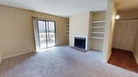 This single-floor residence offers a thoughtfully designed living space that balances comfort and functionality. The main living area serves as the focal point of the home, anchored by a classic brick fireplace framed by a clean white mantel, which adds warmth and character to the room. Flanking the fireplace are built-in shelving units, providing ample display and storage space while contributing to the room's structured, symmetrical aesthetic. The living area is finished with plush wall-to-wall carpeting in a neutral tone that complements the warm beige walls, creating an inviting and cohesive atmosphere throughout. A textured popcorn ceiling adds a traditional architectural detail overhead. Natural light flows generously into the space through a large sliding glass door, which opens to a balcony overlooking a landscaped outdoor area, offering pleasant exterior views and a seamless connection between indoor and outdoor living. The sliding door is dressed with sheer vertical drapes that diffuse the light softly into the room. Overall, the space presents a clean, move-in-ready environment with a warm and neutral palette suited to a variety of interior styles.