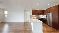 This single-floor residence combines modern design with functional living spaces, finished with a consistent palette of crisp white walls and rich, dark hardwood-style flooring that flows through the main living areas. The open-concept layout connects a generous living room—bathed in natural light from a large multi-pane window—directly to the kitchen and dining areas, creating a seamless, airy atmosphere.

The kitchen is well-appointed with rich espresso-toned cabinetry offering abundant storage, a decorative mosaic tile backsplash, white countertops, and a full suite of stainless steel appliances including a gas range, over-the-range microwave, and a side-by-side refrigerator. A white kitchen island with an integrated sink adds both workspace and functionality.

The bedrooms feature plush carpeting and sliding glass doors that open onto a private balcony, offering expansive outdoor views of the surrounding neighborhood. These rooms provide a calm, comfortable retreat with ample natural light.

Two bathrooms are finished with white subway tile surrounds in the tub/shower combinations, wood-toned flooring, and clean white fixtures. One bathroom includes a rich wood vanity with a white countertop and a large mirror, adding a refined touch to the space overall.