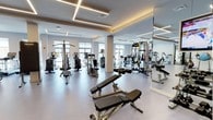 This fitness center offers a comprehensive workout environment designed to accommodate a wide variety of exercise needs. The space features an open, airy layout with a light-toned floor and generously high ceilings adorned with distinctive geometric linear lighting fixtures that provide bright, even illumination throughout. Large windows along the walls allow ample natural light to fill the room, creating an energizing atmosphere for workouts.

The facility is well-stocked with a diverse selection of equipment. A dedicated strength training area includes multi-station cable machines, adjustable weight benches, and a functional trainer, providing options for a full range of resistance exercises. The cardio zone lines one side of the space with a row of treadmills and upright cardio machines such as ellipticals and steppers, ideal for sustained aerobic training. Full-length mirrors are strategically placed along the walls, enhancing the sense of space while allowing users to monitor their form.

The clean, neutral color palette of white walls and light flooring contributes to a modern, professional aesthetic. The thoughtful arrangement of equipment ensures ease of movement and accessibility throughout the floor. Overall, this fitness center delivers a polished and functional environment suited for a comprehensive training regimen.