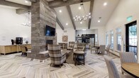 This single-story community clubhouse blends rustic charm with refined elegance through thoughtful architectural detailing throughout. The exterior makes a striking first impression, featuring natural stacked stone facades, bold timber-frame gabled entry, lush landscaped garden beds, and arched transom windows framed by classic coach lanterns.

Inside, the main gathering area is anchored by soaring vaulted ceilings with exposed wood beams and an eye-catching branch-style chandelier that casts warm, inviting light. A floor-to-ceiling stone fireplace serves as the room's dramatic focal point, complemented by plaid upholstered armchairs and herringbone-patterned wood-look flooring. The open layout flows seamlessly into a full kitchen outfitted with dark cabinetry, pendant lighting, stainless steel appliances, and a breakfast bar, alongside a dedicated dining area.

The outdoor spaces are equally impressive. A timber-framed covered patio features a grand stone fireplace, striped lounge seating, and an adjacent outdoor kitchen with a grill. A separate open-air patio area offers a central fire pit surrounded by wicker seating, overlooking a tranquil pond and the natural stone exterior of the main building — ideal for relaxed outdoor gatherings in any season.