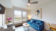 This well-appointed one-bedroom unit offers a thoughtfully designed layout across a single floor, blending contemporary style with practical functionality. The living area serves as the heart of the space, featuring light-toned hardwood-style flooring, a ceiling fan, and large windows that invite natural light and frame views of the surrounding cityscape and greenery. A bold blue sofa anchors the seating area, complemented by a geometric round coffee table and a compact dining set with a wood-top table and vibrant yellow chairs — adding a lively pop of color to the open-plan layout. A striking abstract artwork with gold accents adorns the wall, enhancing the modern aesthetic.

The kitchen is efficiently designed with clean white cabinetry, white countertops, and a full suite of stainless steel appliances, including an electric range, over-the-range microwave, and a sink with ample counter space on either side.

The bedroom is a calm, neutral retreat featuring a ceiling fan, a spacious sliding-door closet, and soft carpeting underfoot. A framed artwork above the bed adds a refined finishing touch.

The bathroom is finished in warm neutral tones, featuring a single vanity with under-sink storage, a lighted mirror, and a shower-tub combo — clean and functional in design.