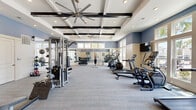 Located at 1290 Kendall Town Blvd in Jacksonville, FL, this well-appointed fitness center offers a generous, open exercise space designed to accommodate a comprehensive workout experience. The room is characterized by striking coffered ceilings with exposed dark wood beams and large ceiling fans that ensure optimal air circulation throughout the space.

Natural light floods the interior through expansive floor-to-ceiling windows positioned along multiple walls, offering pleasant outdoor views of palm trees, greenery, and a nearby pool area. The two-tone wall design — featuring soft blue upper panels above a neutral beige wainscoting — lends the space a clean, modern aesthetic.

The exercise room is comprehensively equipped with a wide variety of fitness machines, including cable and multi-function strength stations, a squat rack, treadmills, ellipticals, stair climbers, stationary bikes, and spin bikes. A full dumbbell rack and additional free weights, along with kettlebells and medicine balls, support diverse strength training routines. Flat-screen TVs are mounted on the walls for entertainment during workouts. Large wall mirrors enhance the sense of depth and allow users to monitor their form. The flooring consists of durable, low-pile carpet throughout, providing a comfortable surface underfoot. The overall ambiance is bright, airy, and motivating.