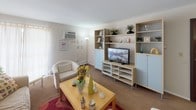 This single-floor residence offers a thoughtfully arranged layout encompassing a living room, bedroom, kitchen, dining area, and full bathroom. The living room serves as the central gathering space, finished with wall-to-wall carpeting and neutral wall tones. It accommodates generous seating and features a modular shelving unit with light blue cabinet doors that houses a flat-screen television, providing ample display and storage space. A wall-mounted air conditioning unit and a window with sheer curtains allow natural light to filter in.

The bedroom continues the neutral palette with carpet flooring and bright white walls. A window dressed in sheer curtains adds natural light, while a closet with a classic wood-paneled door offers convenient storage. The kitchen is a galley-style layout featuring warm wood cabinetry, light countertops, a double-basin sink, and tile flooring, with white appliances including a full-sized refrigerator.

The bathroom includes a single-sink vanity with wood cabinetry, a mirror, and a tub-shower combination finished with white subway tile. The dining area, visible in the overhead view, accommodates a table and chairs, completing this functional and cohesive living space.