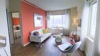 This single-floor space makes a strong visual impression through its thoughtful use of color and contemporary design. The living area is anchored by a bold coral-toned accent wall, which serves as a striking backdrop for a decorative mirror and creates a warm, energetic atmosphere throughout the room. White sheer curtains frame a large window that floods the space with natural light while offering expansive city views, reinforcing a sense of openness despite the intimate scale of the room.

The hardwood flooring in a rich, warm tone runs throughout, grounding the space and complementing the vibrant color palette. Furnishings are modern and curated, featuring a sleek white sofa dressed with colorful throw pillows in green, orange, and blue, adding playful contrast to the neutral upholstery. A round green coffee table acts as a central focal point, paired with sculptural decorative accents that add an artistic quality to the room.

An acrylic side table near the sofa contributes a contemporary, airy feel, while a white swivel chair positioned in the foreground adds both function and visual interest. A secondary seating or console area along the right wall provides additional utility. Overall, the space balances bold design choices with clean, modern lines for a polished, urban aesthetic.