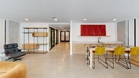Located at 1314 Spring Garden St in Philadelphia's Spring Garden neighborhood, this thoughtfully designed amenity floor offers a well-appointed collection of communal spaces across a single, cohesive level. The layout encompasses six distinct areas connected by a wide, light-filled corridor featuring polished concrete floors, crisp white walls, and black-framed glass partitions that maintain visual openness throughout.

The central lounge serves as the social hub, furnished with a bold amber sectional sofa, a pair of sculptural ivory wingback chairs, and a round pedestal coffee table, all set against floor-to-ceiling windows that frame views of the surrounding Philadelphia streetscape and an adjoining outdoor terrace. A wall-mounted television and striking vertical architectural columns add character to the space.

The adjacent dining area features a long wooden communal table with woven leather chairs beneath dome pendant lights, with large sliding glass doors opening directly to a terrace overlooking neighboring brick architecture. A compact study nook offers a classic leather lounge chair and open wood shelving alongside exposed concrete columns.

The kitchenette is equipped with light wood cabinetry, a stone countertop, and a bar-height seating area, accented by a vibrant red wall installation. The ADA-compliant restroom is clean and functional, finished in polished concrete with grab bar fixtures. Throughout, curated artwork and cohesive modern furnishings reinforce the space's refined, contemporary character.
