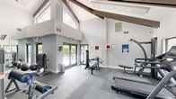 Located in Sunrise, Florida, this fitness center offers a bright and functional space designed to support a comprehensive workout experience. The room's most striking architectural feature is its soaring vaulted ceiling, accented by exposed wood beams that lend a warm, contemporary character to the otherwise clean, modern interior. White-paneled ceiling surfaces and soft gray walls create a fresh, open ambiance throughout.

Natural light floods the space through a generous arrangement of large windows along multiple walls, as well as glass-panel entry doors that provide views of the surrounding landscaped exterior. This connection to the outdoors enhances the airy feel of the room.

The exercise floor is outfitted with durable interlocking rubber flooring tiles, providing both safety and comfort during workouts. Cardio equipment includes treadmills, an elliptical trainer, and a stationary cycle, all positioned to take advantage of the window views. The strength training area features a multi-function cable machine, a leg press, a leg extension/curl machine, a weight bench, and additional selectorized stations, offering a well-rounded selection for resistance training. A wall-mounted mirror further enhances the sense of space. The facility is complemented by safety signage and fire safety equipment, reflecting a commitment to user well-being.