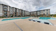 Located at 1004 Diamondback Dr in Sneads Ferry, North Carolina, this outdoor amenity complex offers a premier resort-style pool experience designed for relaxation and recreation. The facility features multiple pool areas, including a large main swimming pool, a dedicated lap lane section, and a separate spa or hot tub, all finished with clean tile detailing and stainless steel entry railings. The pools are set within a generously sized concrete sun deck that wraps around the water features, providing ample space for lounging. Rows of modern navy-cushioned chaise lounge chairs and umbrellas are arranged throughout the deck, offering both shaded and open-air seating options. A dedicated clubhouse or amenity building is positioned along one edge of the complex, featuring large windows and covered outdoor seating, ideal for gatherings or events. The complex is enclosed by a classic black iron fence and is flanked on multiple sides by four-story residential buildings constructed with a mix of brick and neutral-toned siding, featuring arched balconies that overlook the pool area. Mature pine trees frame the perimeter, providing a natural backdrop that enhances the sense of privacy and seclusion. The overall ambiance is that of a well-appointed, community-focused outdoor retreat.