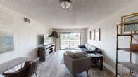This single-floor residence offers a thoughtfully arranged layout that balances comfort and functionality. The living room serves as the centerpiece of the space, featuring wood-look flooring, a textured ceiling, and a large sliding glass door that opens to a private balcony. The balcony provides views of the surrounding greenery and neighboring buildings, allowing natural light to pour into the room. The living area comfortably accommodates a sectional leather sofa, a glass-top coffee table, and a media console, creating a welcoming atmosphere for both relaxation and entertaining.

Adjacent to the living room, the dining area continues the wood-look flooring and is highlighted by a round glass-top dining table with modern white chairs, adding a clean, contemporary touch to the space. A gold-accented shelving unit provides stylish display and storage options nearby.

The bedroom features soft carpeting underfoot, a natural wood platform bed frame, and matching nightstands. A wide window with horizontal blinds invites natural light, while louvered double closet doors offer generous built-in storage. The textured ceiling is consistent throughout the unit, contributing to the cohesive interior character of the space. Overall, the residence presents a clean, modern aesthetic with practical design elements throughout.