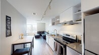 Located on the sixth floor at 24 E Lanvale Street in Baltimore's midtown corridor, this unit offers a clean, modern aesthetic defined by neutral tones, light wood-style flooring, and an efficient single-level layout. The open-concept kitchen and living area serves as the heart of the space, where a streamlined galley kitchen features stainless steel appliances, dark countertops, white cabinetry with open shelving, and a stainless steel sink — all illuminated by adjustable track lighting overhead. A marble-topped dining table with bar-style seating provides a functional transition between the kitchen and living areas. The living room is anchored by large windows that frame expansive views of the Baltimore cityscape, flooding the space with natural light. The bedroom offers a similarly bright atmosphere, benefiting from oversized windows with city and neighborhood views, wood flooring, and a calm, neutral palette conducive to rest. The bathroom is finished with warm earth-toned tile flooring, white subway tile in the shower, and a clean vanity with a dark countertop. Throughout, the unit maintains a cohesive contemporary character with practical finishes and an airy, well-lit ambiance suited to urban living.
